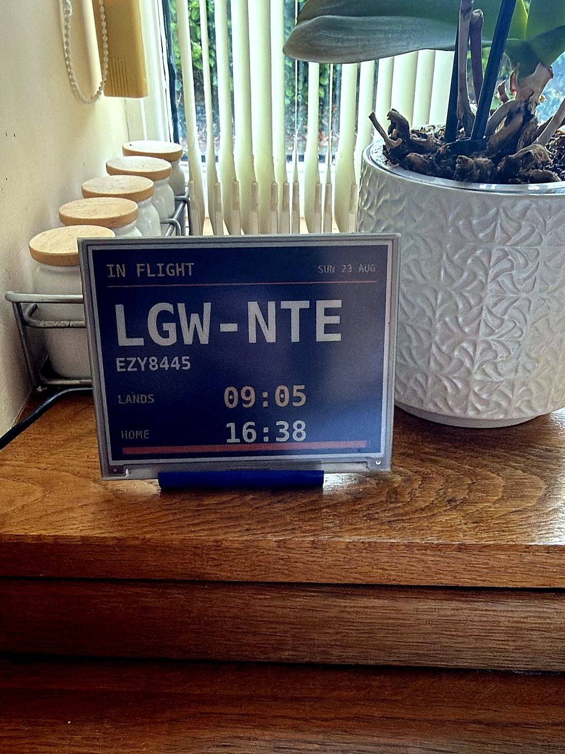 The display on a kitchen worktop beside a plant, showing IN FLIGHT LGW to NTE, landing 09:05 and home 16:38